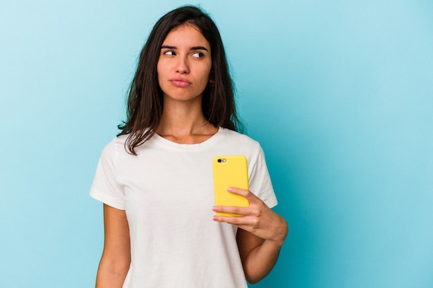Młoda kaukaska kobieta trzymająca telefon komórkowy na białym tle na niebieskim tle jest zdezorientowana, ma wątpliwości i niepewność.