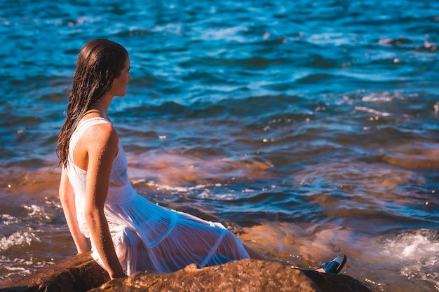 Młoda kaukaska kobieta w mokrej białej sukni, siedząca na skale