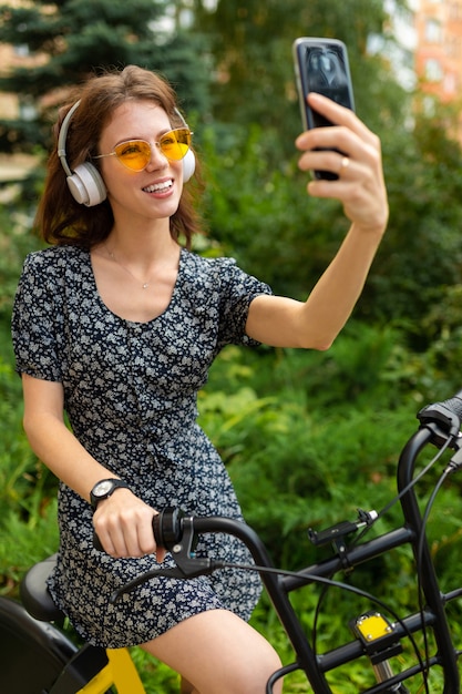 Zdjęcie młoda kaukaska kobieta z doskonałym uśmiechem, pulchne usta, okulary, słuchawki, spacery w przyrodzie, jazda na rowerze i nagrywanie filmów, robienie selfie