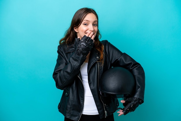 Młoda kaukaska kobieta z kaskiem motocyklowym na białym tle na niebieskim tle szczęśliwa i uśmiechnięta zakrywająca usta dłonią
