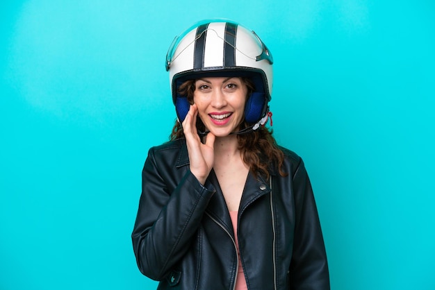 Młoda kaukaska kobieta z kaskiem motocyklowym na białym tle na niebieskim tle ze zdziwieniem i zszokowanym wyrazem twarzy