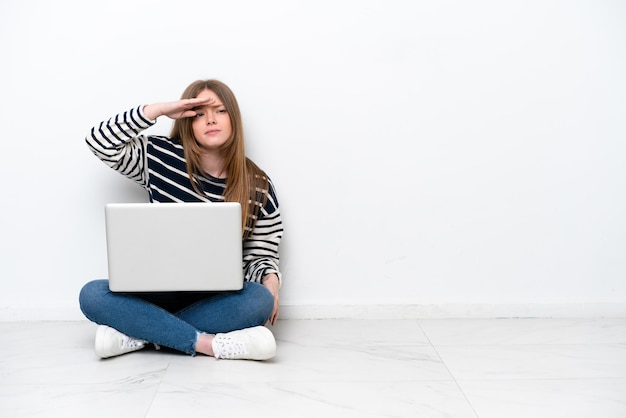 Młoda kaukaska kobieta z laptopem siedząca na podłodze na białym tle patrząca daleko ręką, aby coś wyglądać