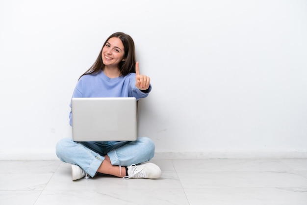 Młoda kaukaska kobieta z laptopem siedząca na podłodze na białym tle pokazująca i unosząca palec