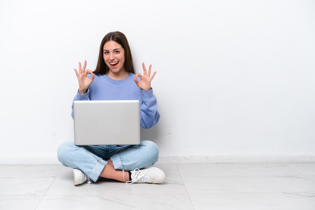 Młoda kaukaska kobieta z laptopem siedząca na podłodze na białym tle pokazująca znak ok palcami