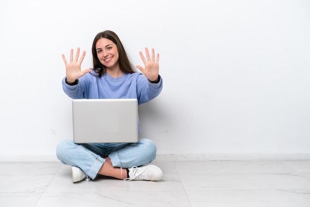 Młoda kaukaska kobieta z laptopem siedzi na podłodze na białym tle, licząc dziesięć palcami