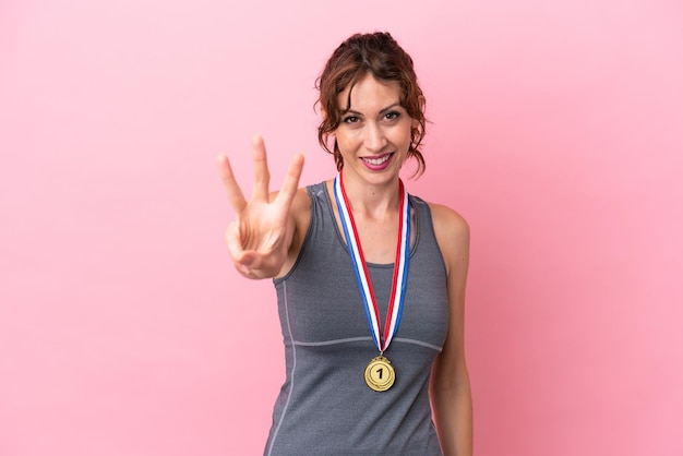 Młoda kaukaska kobieta z medalami na różowym tle szczęśliwa i licząca trzy palcami