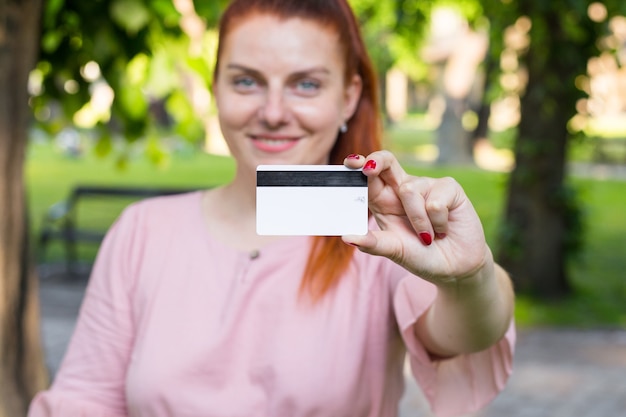 Młoda kaukaska kobieta zostaje w parku i pokazuje białą kartę kredytową z czarną linią magnesu.