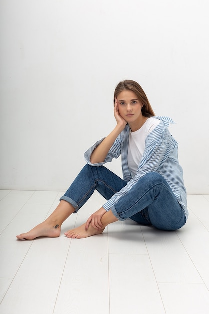 Młoda kaukaska ładna dziewczyna z długimi włosami w koszuli, niebieskie dżinsy siedzi w studio