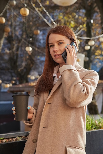 Młoda Kaukaska Rudowłosa Kobieta W Beżowym Płaszczu Z Filiżanką Kawy Dzwoniąca Przez Smartfon Na Zewnątrz