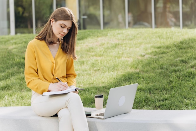 Młoda kaukaska studentka uniwersytetu ucząca się online za pomocą laptopa, robiąca notatki podczas oglądania