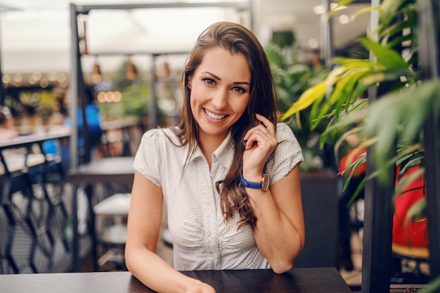 Młoda Kaukaska uśmiechnięta brunetka siedzi w kawiarni i używa mądrze telefon dla połączenia.