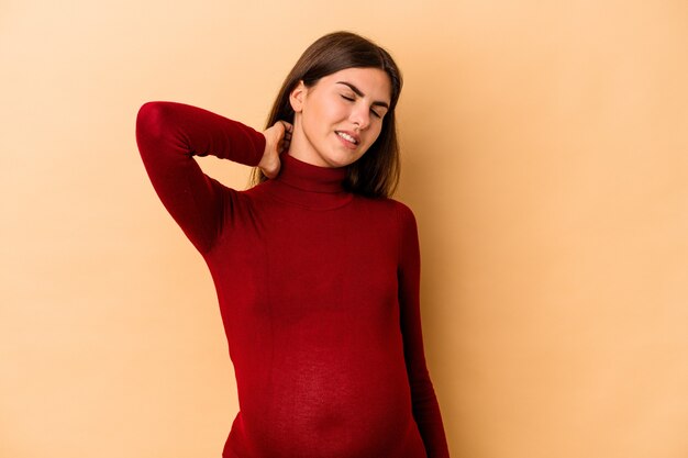 Młoda kaukaski kobieta w ciąży na białym tle na beżowej ścianie cierpi na ból szyi z powodu siedzącego trybu życia.