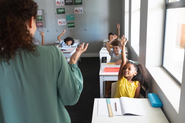 Młoda kaukazyjska nauczycielka ucząca wielorasowych uczniów szkoły podstawowej przy biurku w klasie