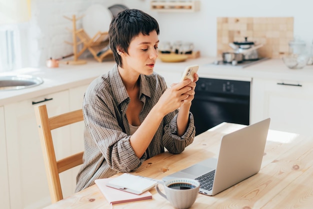 Młoda kaukazyjska współczesna freelancerka siedząca w kuchni używająca telefonu komórkowego i komputera