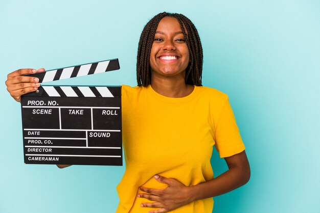 Młoda kobieta african american gospodarstwa clapperboard na białym tle na niebieskim tle, śmiejąc się i zabawę.