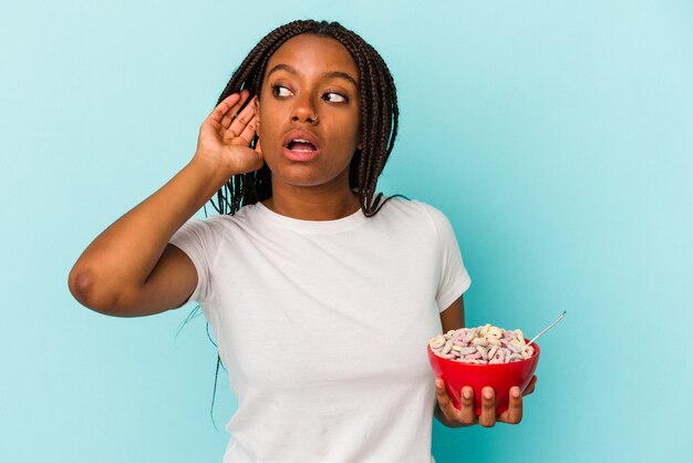 Młoda kobieta Afroamerykanów trzyma miskę zbóż na białym tle na niebieskim tle próbuje słuchać plotek.