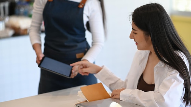 Młoda Kobieta Barista Sobie Fartuch Za Pomocą Tabletu I Odbierz Zamówienie Od Klienta W Kawiarni.