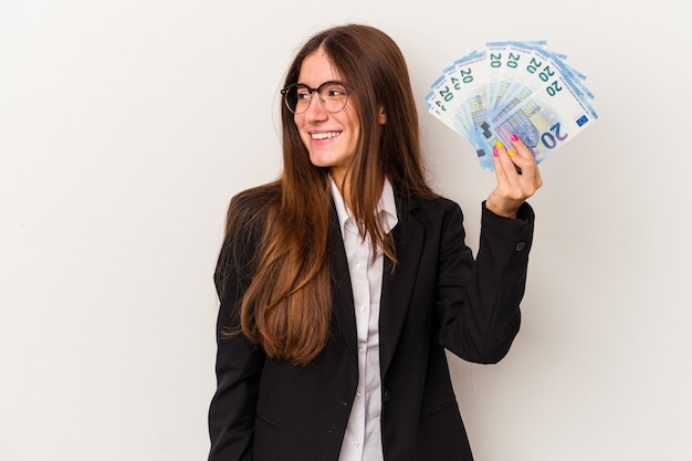Młoda kobieta biznesu kaukaski trzymając banknoty na białym tle wygląda na bok uśmiechnięty, wesoły i przyjemny.