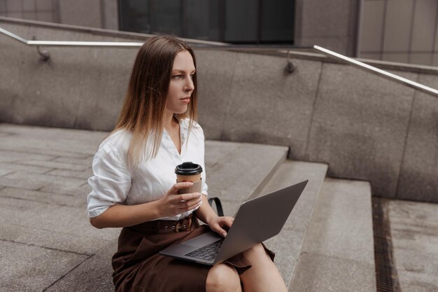 młoda kobieta biznesu trzymająca kawę i laptopa na tle nowoczesnego biurowca