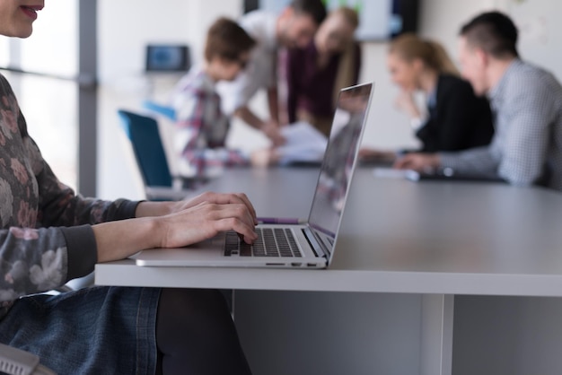 młoda kobieta biznesu w nowoczesnym wnętrzu biura startowego pracująca na laptopie, zespół blured na spotkaniu, grupa ludzi w tle