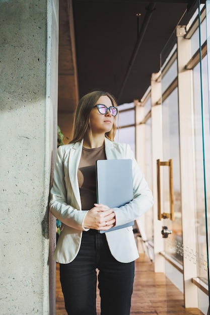 Młoda kobieta biznesu z długimi blond włosami w białej kurtce i okularach stoi z laptopem w dłoniach w przestrzeni biurowej Pracuj w nowoczesnym biurze z dużymi oknami