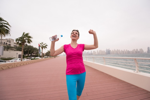 młoda kobieta celebrująca udany trening biegnący promenadą nad morzem z butelką wody i rękami uniesionymi do góry na tle wielkiego miasta