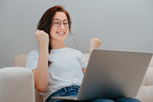 Młoda kobieta cieszy się dobrymi wiadomościami z pracy za pośrednictwem komunikacji wideo online za pośrednictwem laptopa, siedząc w ...