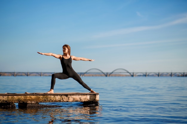 Młoda kobieta ćwiczy ćwiczenia jogi przy cichym drewnianym molo z miastem Sport i rekreacja w pośpiechu miasta