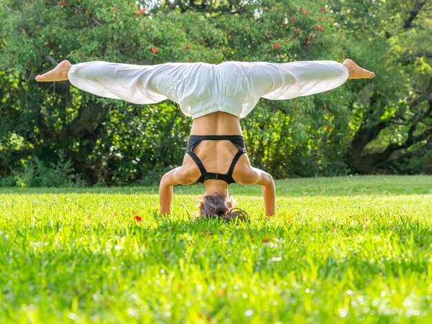 Młoda kobieta ćwiczy jogę stojąc na głowie z rozłożonymi nogami w zielonej koncepcji ParkWellness