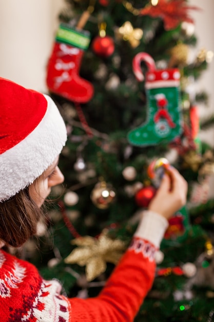 Młoda kobieta dekorowanie choinki z laskami cukrowymi i zabawkami w domu, ubrana w Santa hat. Przygotowanie do Nowego Roku.