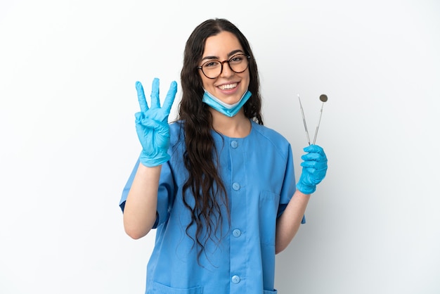 Młoda kobieta dentysta gospodarstwa narzędzia na białym tle szczęśliwa i licząc trzy palcami