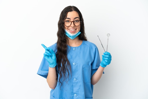 Młoda kobieta dentysta trzyma narzędzia izolowane na białym tle wskazując na bok, aby zaprezentować produkt