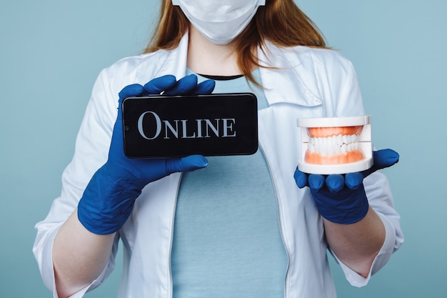 Młoda kobieta dentysta w masce i fartuchu, trzymając instrumenty i telefon. Spotkanie online cocnept.
