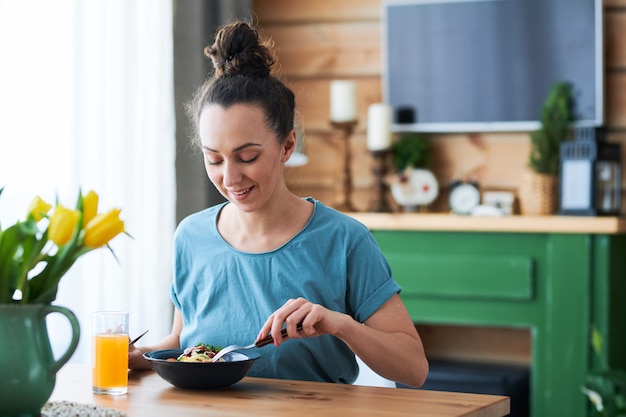 Młoda kobieta dorywczo siedzi przy stole w kuchni podczas jedzenia spaghetti i picia soku owocowego