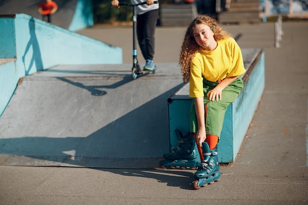 Młoda kobieta dziewczyna w zielonych i żółtych ubraniach i pomarańczowych pończochach z kręconymi fryzurami na rolkach siedząca w skate parku