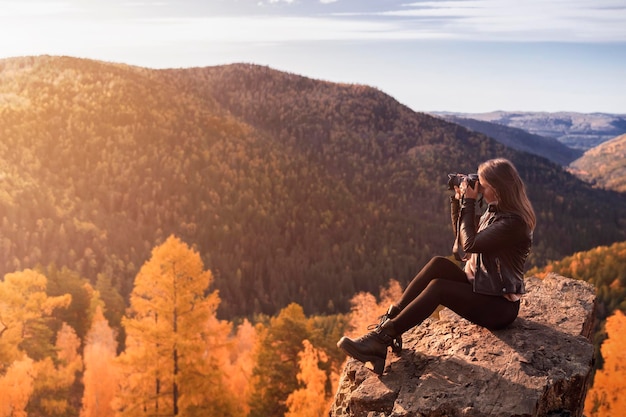 Młoda kobieta fotografuje piękny krajobraz wieczorem siedząc na skale na szczycie góry zmieniając pory roku piękną przyrodę i fotografię hobbystyczną