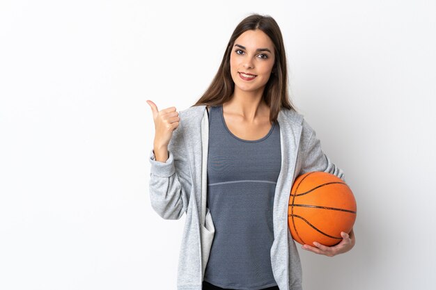 Młoda kobieta gra w koszykówkę na białym tle na białej ścianie, wskazując na bok, aby przedstawić produkt