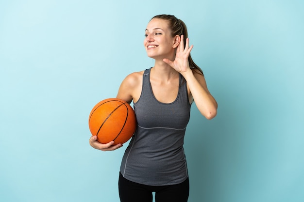 Młoda kobieta gra w koszykówkę na białym tle na niebieskim tle słuchając czegoś, kładąc rękę na uchu