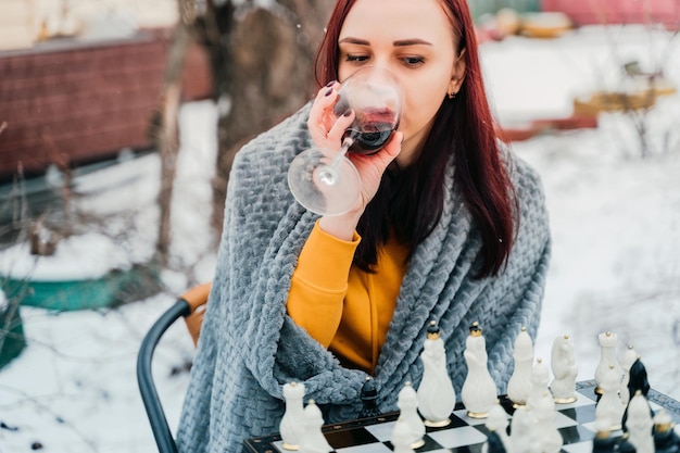 Młoda kobieta gra w szachy i pije czerwone wino na podwórku Kobieta owinięta w szarą kratę z alkoholem siedzi na ulicy grając w grę planszową w sezonie zimowym