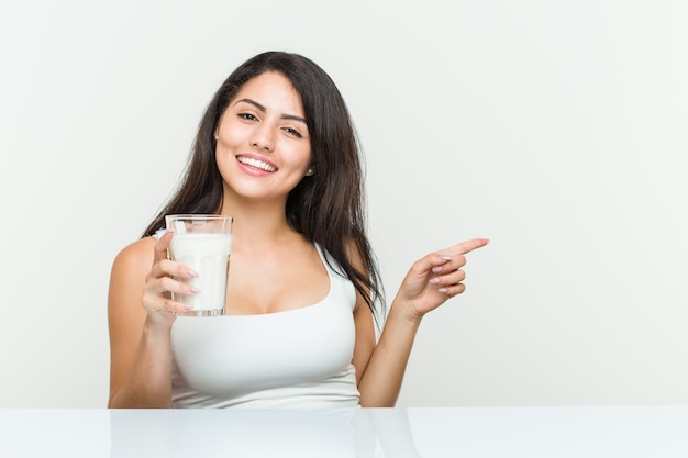 Młoda kobieta hiszpanin trzyma szklankę mleka uśmiechnięty radośnie, wskazując palcem wskazującym z dala.