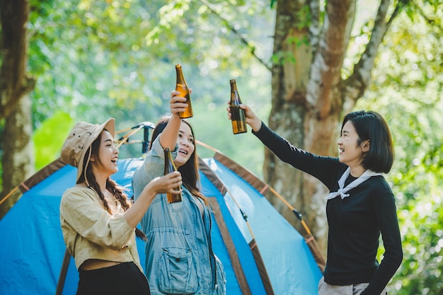 Młoda kobieta i przyjaciółki podróżni relaksujący się na krzesłach kempingowych w namiocie Dopingują i piją piwo podczas kempingu, rozmawiając z zabawą i szczęśliwi razem