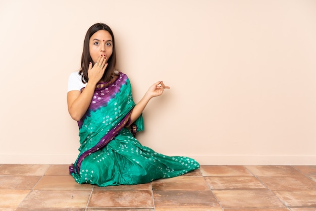 Młoda kobieta indyjska siedzi na podłodze z wyrazem zaskoczenia, wskazując jednocześnie stronę