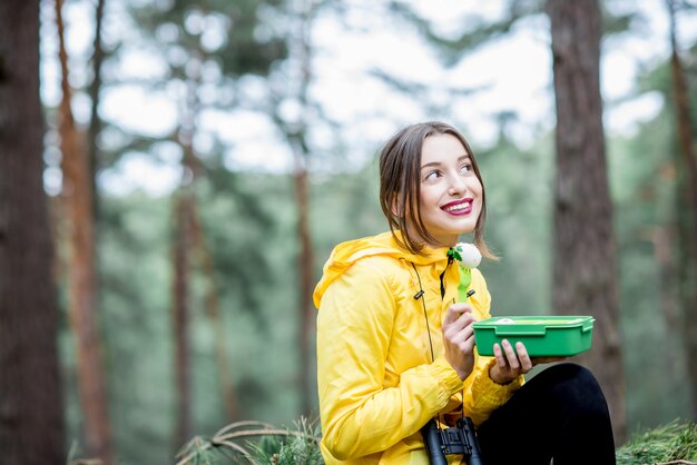 Młoda kobieta je przekąskę ze zdrową żywnością w pudełku na lunch podczas podróży po lesie