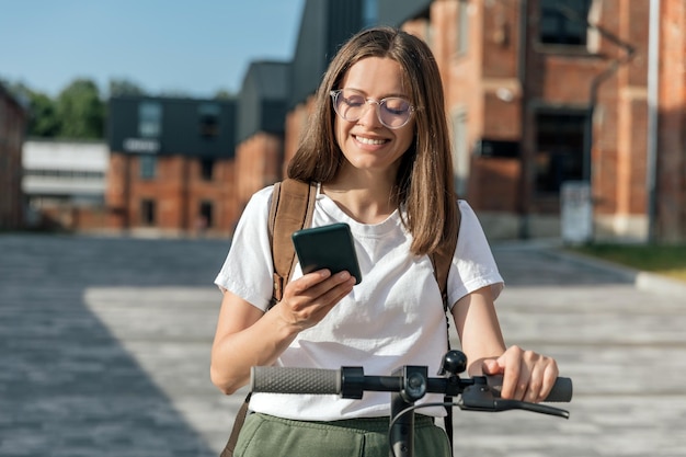 Młoda kobieta jedzie na skuterze elektrycznym i trzyma smartfon w ręku na miejskiej ulicy