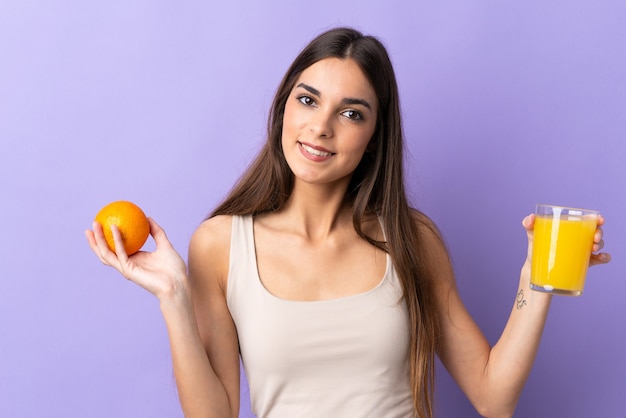 Młoda kobieta kaukaska na białym tle na fioletowej ścianie trzyma sok pomarańczowy i pomarańczowy