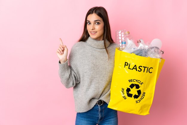 Młoda kobieta kaukaska trzyma torbę pełną plastikowych butelek do recyklingu na białym tle na różowym tle, wskazując na bok, aby przedstawić produkt