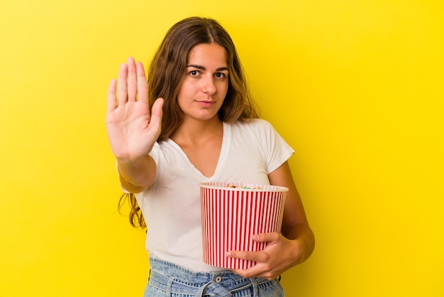 Młoda kobieta kaukaski gospodarstwa popcorns na białym tle na żółtym tle stojąc z wyciągniętą ręką pokazując znak stop, uniemożliwiając.