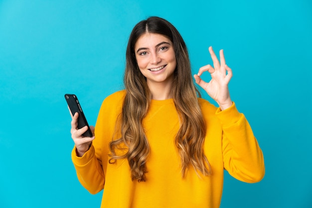 Młoda kobieta kaukaski na białym tle na niebieskiej ścianie przy użyciu telefonu komórkowego i robi znak OK