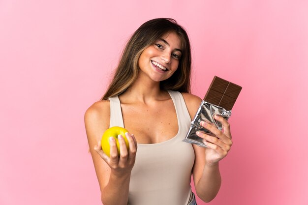 Młoda kobieta kaukaski na białym tle na różowej ścianie, biorąc tabletkę czekolady w jednej ręce i jabłko w drugiej