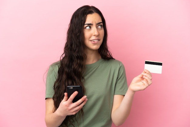 Młoda kobieta kaukaski na białym tle na różowym tle kupowanie z telefonem komórkowym kartą kredytową podczas myślenia